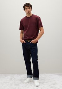 Camiseta de manga corta color marrón con un bolsillo en el pecho, combinada con jeans azul oscuro y zapatillas blancas. Tejido suave con un corte relajado.