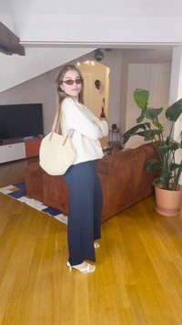 Hvit strikkegenser, mørkeblå vide bukser, hvite hælsandaler, vevd kremfarget skulderveske og brun kordfløyelssofa i bakgrunnen.