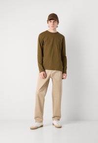 Olivenfarbendes Langarmhemd mit kleinem Logo, beigen Hose und weißen Turnschuhen mit Gummisohlen. Das Modell trägt außerdem eine passende Mütze.