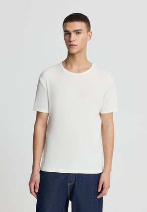 Giovane uomo con capelli castani corti che indossa una maglietta bianca semplice a maniche corte e jeans blu scuro, in piedi davanti a uno sfondo grigio chiaro.
