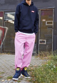 Persona de pie al aire libre, vistiendo una sudadera azul marino, pantalones rosa claro y zapatillas de baloncesto Ewing azul marino con amarillo, cerca de una puerta metálica.