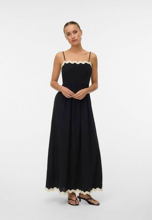 Mujer de pie con un vestido maxi negro de tirantes finos y ribete beige ondulado en el borde superior y el dobladillo, combinado con sandalias negras de tacón.
