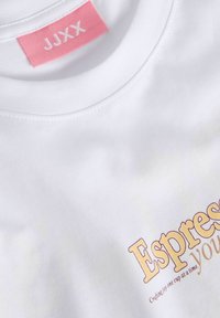 Biały t-shirt z różową metką JJXX wewnątrz kołnierzyka oraz częściowym żółto-brązowym napisem "Espres" i mniejszym tekstem poniżej.