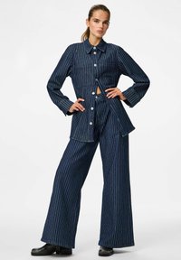 Denim apģērbs, kas sastāv no pogājama žaketes un plašiem biksēm, abiem ar tumši zilu un baltu vertikālu svītru rakstu. Mīksta tekstūra un brīva piegriezuma.