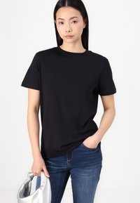 Black cotton t-shirt with short sleeves and a crew neckline, paired with blue jeans. Model holds a silver bag in one hand.
