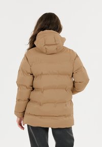 Person med langt brunt hår iført en tan farvet pufferjakke med hætte, står mod en plain hvid baggrund, vendt væk.
