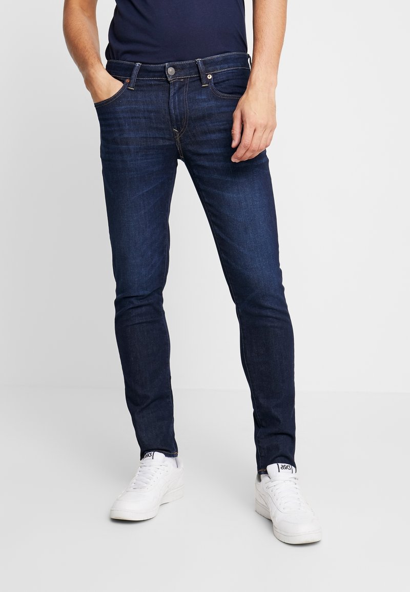 Homme portant un jean slim bleu foncé et des baskets blanches, debout avec une main dans la poche sur un fond clair uni.