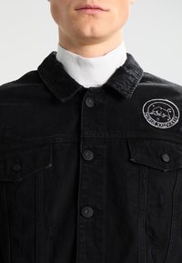 Zwarte spijkerjack met een knoopsluiting aan de voorkant, twee borstzakken en een geborduurd rond logo. Versleten textuur voegt detail toe.