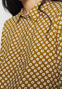 Camicia button-up marrone con un motivo floreale caratterizzato da fiori bianchi con centri neri, realizzata in un tessuto texture.