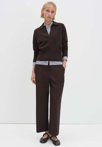 Brauner Strickpullover mit V-Ausschnitt, darunter ein gestreiftes Hemd mit Kragen, und braune, weit geschnittene Hosen. Schwarze flache Schuhe runden das Outfit ab.