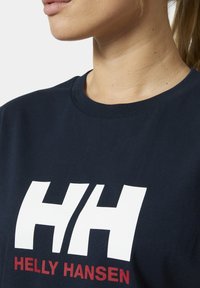 Primer plano del cuello y la parte superior del pecho con una camiseta azul marino que tiene un gran logo blanco "H/H" y el logo rojo "Helly Hansen".