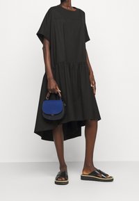 Woman wearing a loose black high-low dress with short sleeves, holding a blue handbag, and wearing black platform slide sandals.