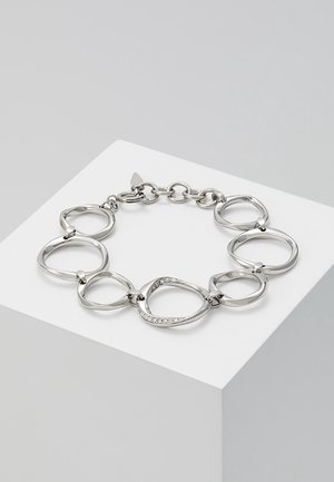 Silbernes Kettenarmband mit unregelmäßigen ovalen Gliedern, ein Glied mit kleinen klaren Kristallen besetzt, auf einer weißen Plattform präsentiert.