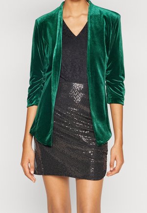 Persoon draagt een groen fluwelen blazer over een zwarte kanten top en een zwarte minirok met pailletten, staand tegen een effen achtergrond.