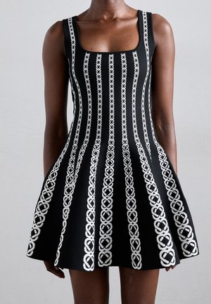 Schwarzes ärmelloses Kleid mit weißen vertikalen kettenartigen Streifen, getragen von einer Person vor einem einfarbigen hellen Hintergrund.
