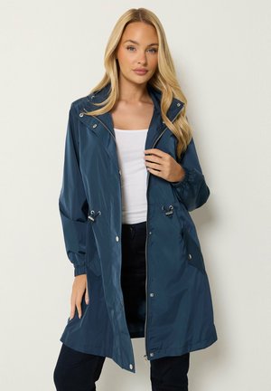 Femme aux longs cheveux blonds portant un imperméable bleu marine par-dessus un haut blanc et un pantalon noir, debout devant un fond clair.