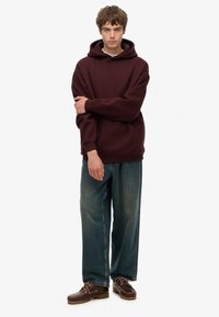 Sudadera burdeos con capucha y corte holgado, combinada con jeans azules sueltos y zapatos marrones con cordones. Textura suave en toda la prenda.