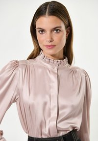 Langärmlige Bluse aus zartem rosa Satin mit Rüscheneinsatz am hohen Kragen, plissierten Schultern und Knopfleiste, kombiniert mit dunkelblauer Jeans.