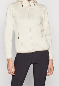 Frau in einer strukturierten cremefarbenen zip-up Kapuzenjacke mit Kordelzügen und schwarzen Leggings, die vor einem schlichten Hintergrund steht.