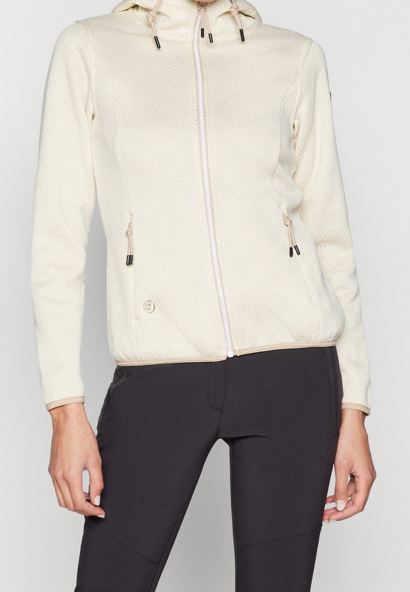 Frau in einer strukturierten cremefarbenen zip-up Kapuzenjacke mit Kordelzügen und schwarzen Leggings, die vor einem schlichten Hintergrund steht.