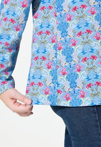 Blaues Blumenhemd mit rosa und gelben Blumen, runder Saum und geknöpften Manschetten. Weicher Stoff mit filigranen botanischen Mustern.