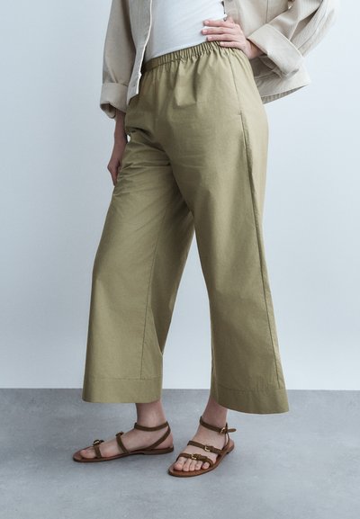 Personne portant un pantalon large court couleur olive claire avec taille élastique et sandales plates marron, debout sur un sol gris devant un fond pâle.