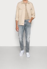 Chaqueta ligera beige con dos bolsillos en el pecho, camiseta color trigo, jeans azul claro y zapatillas blancas, de pie frente a un fondo liso.