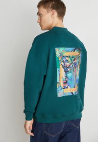 Teal sweatshirt met een kleurrijke grafische print op de achterkant, die abstracte gezichten, bladeren en druiven toont. Casual pasvorm, geribde manchetten.