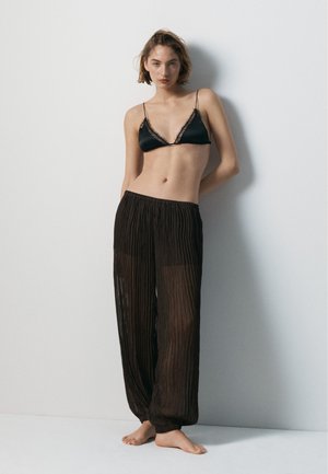 Kobieta stoi boso przy ścianie, ubrana w czarny bralette z koronkowym wykończeniem oraz przezroczyste, plisowane czarne spodnie o szerokich nogawkach.