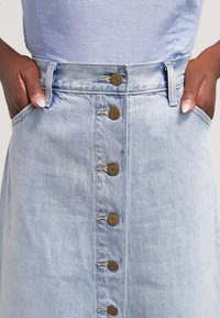 Hellblauer Jeansrock mit Knopfleiste, ausgestattet mit bronzefarbenen Knöpfen und Seitentaschen, aus weichem, strukturiertem Stoff gefertigt.