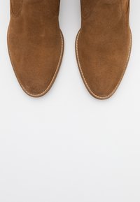 Bruna mockakängor med spetsig tå, med synliga sömmar längs sulan och en slät textur. Inga märkbara mönster eller detaljer.