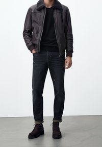 Brun läderbomberjacka med fårskinnskrave, svart tröja, mörka denimjeans och svarta snörskor. Slät textur och figursydd form.