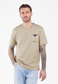 Hombre con tatuajes que lleva una camiseta beige con una paloma y el texto "ESPERO", jeans de un azul claro, de pie con una mano en el bolsillo sobre un fondo blanco.