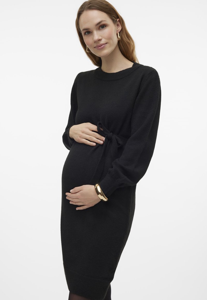 Black knit maternity dress with a crew neckline, long sleeves, and a tie waist. Smooth texture and fitted shape, reaching mid-thigh.