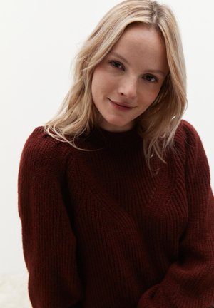 Burgunderfarbener, gestrickter Pullover mit hohem Kragen, der ein strukturiertes Muster und Rippdesign aufweist. Das Modell hat hellbraunes, sanft umrahmtes Haar.