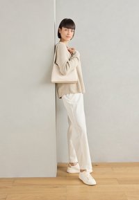 Un pull en tricot beige, un pantalon blanc côtelé et des baskets blanches. Un petit sac à main beige repose sur l'épaule. Contre un mur neutre.