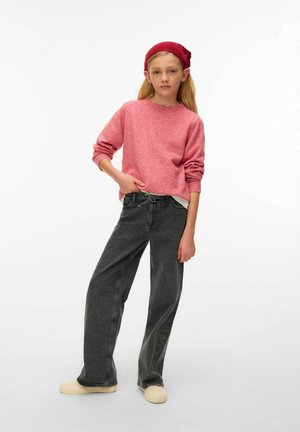 Růžový pletený svetr s dlouhými rukávy, doplněný černými džíny s vysokým pasem a béžovými nazouvacími botami. Modelka pózuje s jednou rukou v pase.