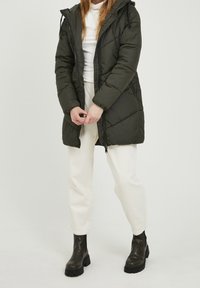 Žena v tmavě zeleném prošívaném kabátu, bílém roláku, bílých kalhotách a tmavě zelených robustních kotníkových botách stojí na bílém pozadí.