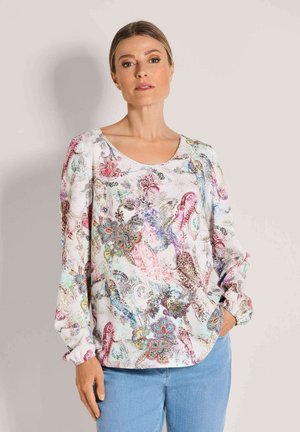 Frau trägt eine langärmelige Bluse mit floralem Muster und rundem Ausschnitt sowie hellblaue Jeans, steht mit einer Hand in der Tasche.