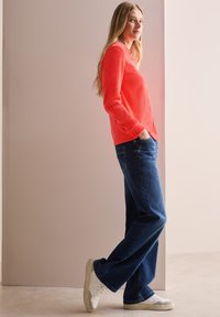 Koraalkleurig shirt met lange mouwen van gestructureerde stof, gecombineerd met donkerblauwe wijde jeans en witte sneakers, staande voor een neutrale achtergrond.