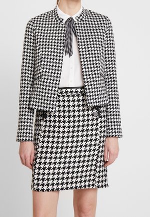Vrouw draagt een zwart-wit pied-de-poule patroon jasje en bijpassende rok over een witte blouse met een klein zwart-wit strikje.