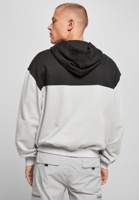 Uomo con capelli corti che indossa una felpa a blocchi di colore bianco e nero e pantaloni cargo grigi chiari, rivolto a sinistra su uno sfondo grigio.