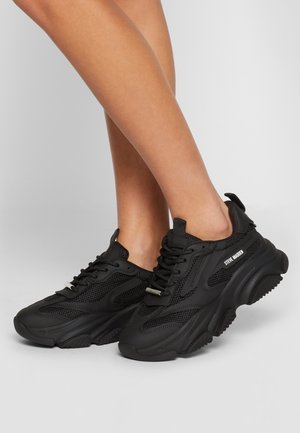 Jambes portant des baskets noires Steve Madden à semelles épaisses et panneaux en mesh, posées devant un fond gris clair uni.