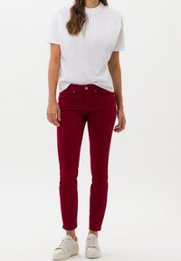 Pantalones de algodón rojos con un ajuste slim, diseño de cinco bolsillos y cierre de un solo botón. Combinados con una blusa blanca de manga corta y zapatillas blancas.