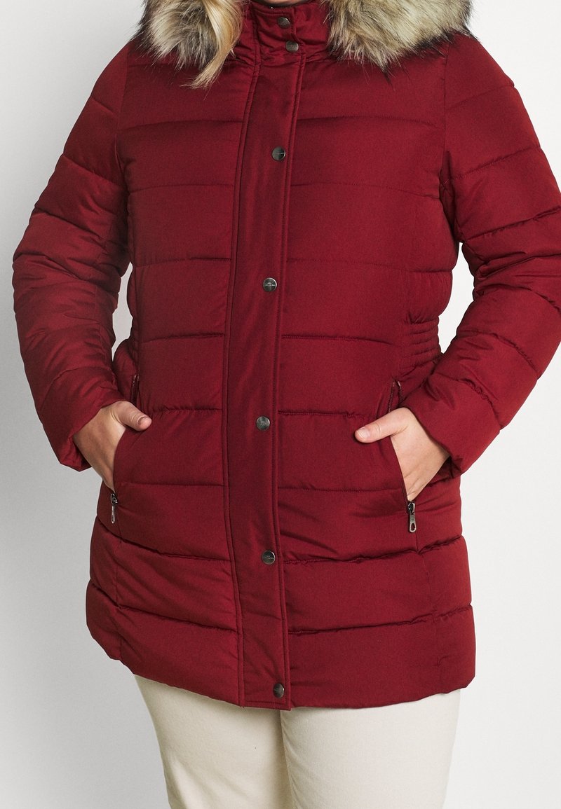 Burgundy pufferjacka med en huva av konstgjord päls, har horisontell sömnad, dragkedja fram och sidofickor med dragkedjestängning.