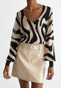 Maglione nero e beige a V con motivi ondulati, maniche lunghe e tessuto lucido. Abbinato a una mini gonna in pelle beige con cintura.