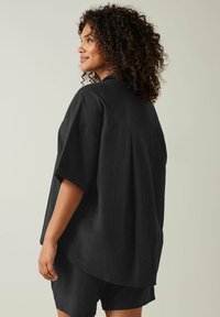 Maglietta nera, larga, realizzata in tessuto testurizzato con maniche corte, orlo arrotondato e un design della spalla posteriore dritto. Abbinata a pantaloni corti.