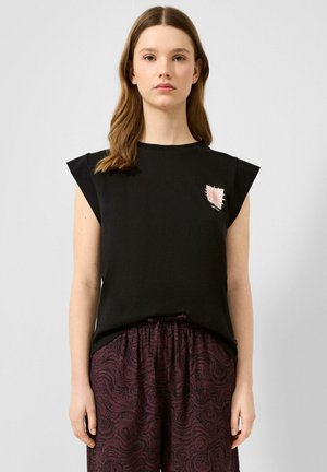 Jeune femme aux cheveux mi-longs portant un haut noir sans manches avec un motif rose sur la poitrine et un pantalon bordeaux à motifs.
