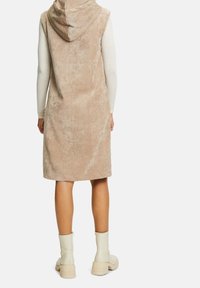 Kvinna som bär en beige huva utan ärmar klänning över en långärmad tröja och vita ankelskor, stående vänd bort från kameran mot en vit bakgrund.