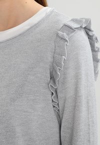 Silverstickad tröja med rynkade detaljer på axlarna, med en mjuk textur och lätt lyster, lagd över en vit skjorta.